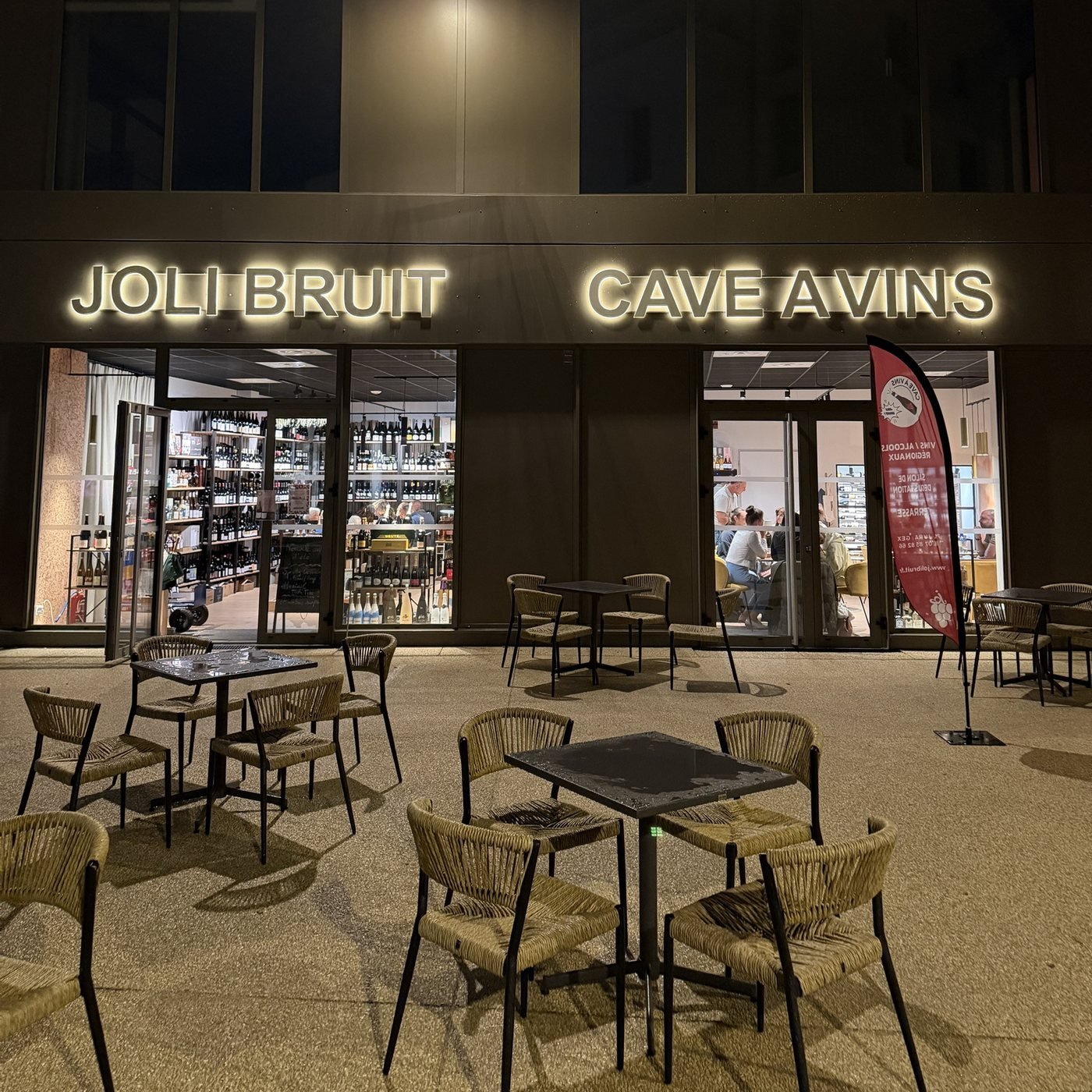 Devanture illuminée et terrasse de Joli Bruit, cave à vins à Gex, en soirée